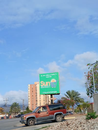 The image depicts a bright, sunny day in Juan Griego, Venezuela. The sky is a clear blue with scattered white clouds. In the foreground, a rusty brown Chevrolet SUV is parked on the side of a road, with its driver, a young female with light-colored hair, visible behind the steering wheel. The SUV is facing to the left. Behind the SUV, a tall, multi-story building with many windows is visible, colored in shades of orange and beige. To the right of the building, a large green billboard is prominently displayed, advertising "Sun Market" with the slogan "EL SOL BRILLA PARA TI, AQUI EN". A shopping cart icon is depicted on the billboard. Further to the right, a street lamp is adorned with red flowers. The environment appears to be urban, with palm trees and other vegetation visible. The ground is covered with gravel and debris in the immediate foreground. Other vehicles and buildings are visible in the background, suggesting a bustling city scene.