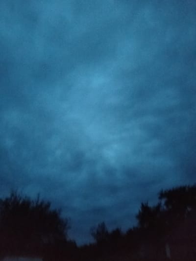 The image depicts a dark, overcast sky with indistinct cloud formations, rendered in various shades of blue and grey. The lower portion of the image features the silhouettes of trees, appearing as dark masses against the dimly lit sky. There are no people, specific structures, or identifiable landmarks visible in the frame. The overall atmosphere is moody and subdued, suggesting either twilight or pre-dawn conditions. The location provided, Ustyvytsia, Ukraine, is not discernible from the visual elements of the photograph.