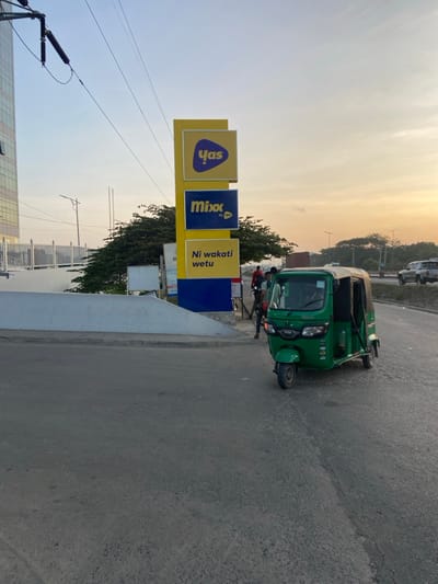 The image shows a green three-wheeled auto-rickshaw parked on the side of a road in Dar es Salaam, Tanzania, at the coordinates -6.779, 39.218. Two people are standing nearby. The auto-rickshaw is facing towards the camera. In the background, there are power lines, street signs, and a building. One sign advertises the words "Mix" and "Yas" in white on a yellow background. Another sign is in Swahili and reads "Ni wakati wetu." There are trees on both sides of the street. The sky is a light blue with a few white clouds and the sun is setting in the distance. The road is paved with asphalt and appears to be in good condition. The environment is a typical urban scene.