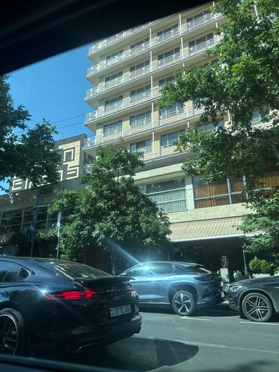 The image shows a street scene in Yerevan, Armenia. In the foreground, a black Porsche Panamera is parked on the right side of the road. To its right, a blue electric SUV is parked, and further to the right, a dark-colored Mercedes-Benz is partially visible. In the background, a multi-story building with balconies is prominent. The building appears to be commercial or residential, with windows and balconies visible on each floor. Trees with green foliage line the street, providing shade. The sky is clear and blue. The building on the left side of the image has large windows and an awning. A sign with Cyrillic lettering, "ЕВРОПА" (Europe), is visible on this building. The license plate on the Porsche is from Armenia, displaying "37 TE 436". The overall scene suggests a typical urban environment with parked cars and a mix of modern and older architecture.