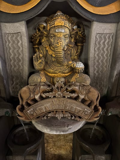 The image shows a close-up, overhead view of a decorative fountain sculpture, likely at a temple or religious site in Kuta, Indonesia. The central subject is a golden statue of Ganesha, the elephant-headed Hindu deity, depicted in a seated position. Ganesha is adorned with intricate jewelry and clothing, with a prominent forehead marking.
Surrounding Ganesha is a carved wooden or stone embellishment featuring a shield-like element with the word "GALLERY" carved into it. The shield is supported by what appears to be carved lions or other mythical creatures. Water is actively spraying from spouts integrated into the sculpture, flowing into dark, circular basins below. The sculpture is set against a backdrop of concrete or stone architectural elements, some with a wave-like pattern. The lighting suggests it might be indoors or under covered outdoor conditions, with no clear indication of time of day or weather. The overall impression is one of a detailed and ornate religious or cultural art installation.