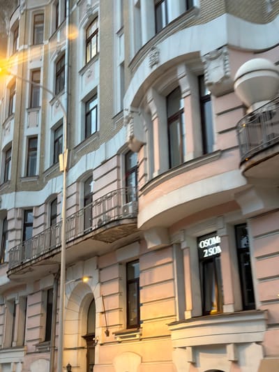 This is a daytime, exterior, low-angle, full shot of a building in Moscow, Russia. The building appears to be old and is made of brick with decorative elements. It has many windows, some of which are arched, and balconies with ornate railings. There is a street lamp in the foreground, and a sign with text is visible on one of the windows. The building is painted in light colors, with pink and beige accents. The overall impression is one of aged elegance.