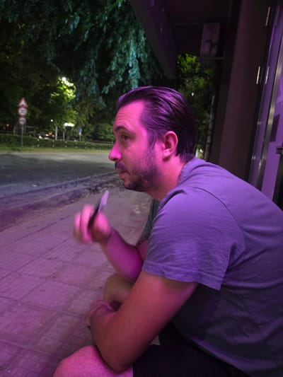 A man is sitting on a sidewalk at night, in front of a building with a dark facade. He is looking to his left, with his right hand raised, holding a small object. The lighting is predominantly purple, casting an unusual hue on the scene. In the background, trees and a street with a speed limit sign are visible. The man has short, dark hair, a beard, and is wearing a purple t-shirt. The sidewalk appears to be paved with square tiles.
