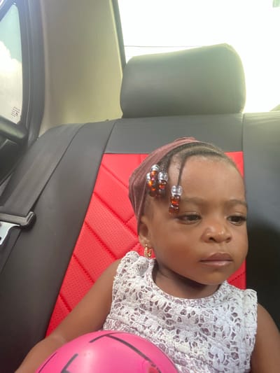 A young child is seated in the rear passenger seat of a vehicle in Adankolo, Nigeria. The child, with dark skin and dark eyes, maintains a neutral expression, gazing slightly to the right of the frame. They wear a dark reddish-brown headwrap, beneath which braided hair with clear and amber beads is visible on the forehead. Small gold-colored hoop earrings are present. The child is dressed in a light-colored, sleeveless top with a distinctive lace or crocheted pattern. A bright pink ball rests in the child's lap. The vehicle's seat features black upholstery with a red, perforated, quilted central insert. A black headrest is positioned behind the child, and a black seatbelt is visible on the left side of the frame. Bright daylight illuminates the scene, visible through the vehicle's window.