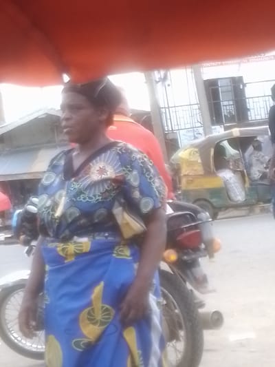 In Gwong, Nigeria, a woman stands in front of a motorcycle, her body turned slightly to the left. She is wearing a vibrant blue and yellow patterned dress, with the fabric of her skirt featuring large yellow circular motifs. A smaller yellow bag is tucked into her waist. The background shows a street scene, with a tuk-tuk vehicle visible to the right of the woman. The tuk-tuk is primarily yellow and green, with its doors open. In the distance, there appears to be a building with a metal gate or fence. The overall lighting suggests it is daytime, likely overcast or hazy. The woman's expression is neutral. Visible text on a sign in the background reads "CONTACT: 07066605 34, 09044426879". Another partial text on a sign beneath the building reads "ONYIAZU".