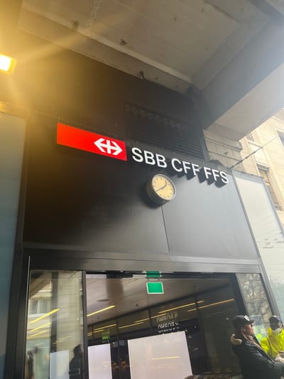 The image depicts an exterior view of a building entrance, likely a railway station, in Geneva, Switzerland. The scene is dominated by a large, dark, rectangular panel serving as a sign or facade element.
On this dark panel, prominent white capital letters spell "SBB CFF FFS", identifying it as a facility of the Swiss Federal Railways. Below the text is the distinct red SBB logo, featuring two white arrows pointing horizontally outwards from a central point. To the right of the text, a round, cream-colored analog clock with black hands and numerals is mounted, indicating a time of approximately 10:10.
Adjacent to the dark panel is a glass facade, possibly an entrance. This glass reflects the interior of the building, revealing horizontal strip lighting, dark walls, and what appear to be light panels or display screens. Through these reflections, two individuals are visible. One person, positioned in the upper right, wears a high-visibility yellow or green jacket, possibly indicating staff or a worker. Another person, located slightly lower, is seen wearing a dark baseball cap and a dark jacket.
The surrounding architecture, including a concrete or stone building section above and to the left, suggests an urban setting. The lighting is bright and diffused, indicating daytime, possibly under an overcast sky or within a brightly lit covered area. A bright yellow light source is visible in the bottom-left corner of the image. The visible text confirms the location's association with the Swiss national railway system. No specific activity beyond people being near an entrance is clearly discernible, but it implies entry or exit from a transportation hub.