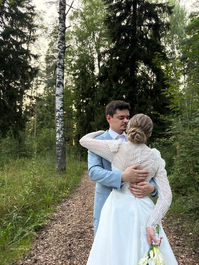 In the city of Bakhtiyary, Russia, a wedding couple embraces on a path in a wooded area. The groom, wearing a light blue plaid suit with a white shirt and no tie, has his arms around the bride. The bride is wearing a white wedding dress with a beaded bodice and long sleeves, and she is holding a bouquet of white flowers. The path is covered in wood chips, and tall green grass and trees surround the couple. The trees are a mix of deciduous and coniferous, with a notable birch tree standing tall to the left of the couple. The sky is overcast, and the lighting suggests it may be late afternoon or early evening. The couple's bodies are close, with their faces touching. A wedding ring is visible on the groom's left hand. The overall atmosphere is intimate and romantic.