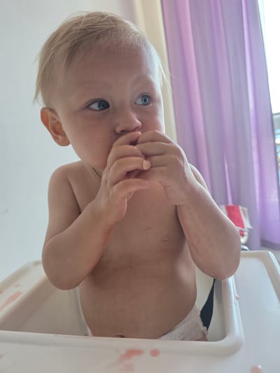 A young, fair-haired child, wearing only a white diaper, sits in a white high chair. The child has a light complexion and blue eyes, and is looking to the right with their hands near their mouth. The high chair has red smudges on its tray. In the background, sheer purple curtains hang over a window. The scene appears to be indoors.