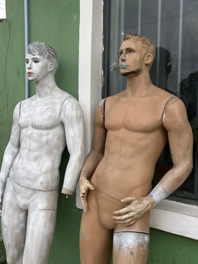 Two weathered male display mannequins are positioned against a building exterior in Lekki, Nigeria. The mannequin on the left is painted in a textured, off-white or light grey, with sculpted grey hair details and orange lipstick. Its lower arms are missing, and it leans against a faded, cracked light green wall. The mannequin on the right, predominantly tan-colored, shows extensive surface wear, grime, and discoloration. It features blue paint smeared around its mouth and chin, and its forearms and hands are a dirtier beige. Its hands are positioned at its groin, holding a thin, light-colored wire. This mannequin is missing its lower legs from just above the knees and leans against a white vertical architectural trim beside a dark, reflective window.