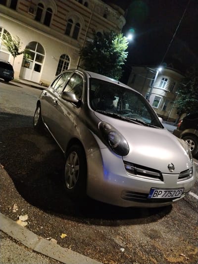 A silver Nissan Micra is parked on a street in Vratsa, Bulgaria at night. The car is positioned at an angle, facing towards the right side of the frame. The vehicle's headlights and taillights are not illuminated. The car's license plate is visible and reads "BP 7752 CP". The street is paved with asphalt and appears wet, with fallen leaves scattered on the ground. A concrete curb runs along the edge of the street. In the background, there is a building with arched windows and decorative horizontal bands. Trees with green foliage are also visible, illuminated by streetlights. Another dark-colored vehicle is parked behind the Nissan. The scene is lit by streetlights and ambient light from the surroundings.