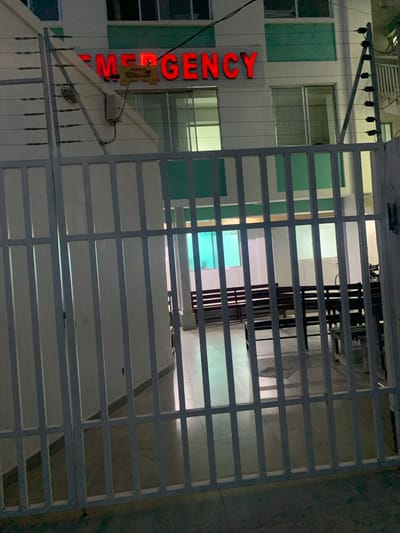 The image shows a partially obscured view of an emergency room entrance through a white metal gate. The emergency sign is illuminated in red vertical letters. The building behind the gate appears to be a multi-story structure with a white facade and green window accents. Inside, seating arrangements are visible. The location of the building is Dar es-Salaam, Tanzania.