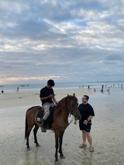This is a vertical, eye-level, medium shot of a beach scene in Kuta Selatan, Indonesia, with two men and a horse as the primary subjects, and a cloudy sky, ocean, and distant shoreline in the background. The man on the left is seated on a brown horse, facing right, and looking down at his phone, which he holds in his right hand. He is wearing a black t-shirt, gray cargo pants, and black boots. The horse is facing right and has a black saddle with a white inscription on its side, and a black bridle. The man on the right is standing to the right of the horse, facing left, and looking at the horse. He is wearing a black t-shirt, black shorts, and tan sandals. His left hand is holding a phone, and he has a smartwatch on his right wrist. The beach is wet, with shallow water reflecting the overcast sky. In the distance, the ocean meets the shoreline, with several traditional Indonesian fishing boats visible on the water. Other people are scattered along the beach and in the shallow water, some walking, some sitting, and some wading. The sky is filled with a dense layer of gray and white clouds.