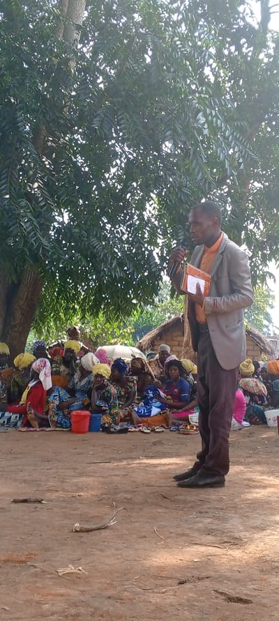 An outdoor scene in Fizi, Democratic Republic of the Congo, depicts a male speaker addressing a large group of seated women. The speaker, positioned on the right side of the frame, is a light-skinned male with short dark hair, wearing a light grey blazer over an orange collared shirt and dark trousers, with dark shoes. He holds a black microphone to his mouth with his right hand and a small, worn, orange-brown notebook or book in his left hand, facing towards the left.
The audience consists of approximately 20-30 women, predominantly of dark skin tone, seated on the dirt ground across the mid-ground and left side of the image. They are dressed in various brightly patterned fabrics, many wearing distinct head coverings in yellow, white, pink, and blue. Several women have plastic buckets (red and blue) positioned near them on the ground. A woman near the center of the seated group holds a baby wrapped in a blue and white blanket and wears a blue and white patterned shirt. Multiple pairs of sandals are visible on the ground in front of the seated individuals.
The environment features a large, leafy tree canopy overhead in the upper left and center, casting shadows. The trunk of this tree is visible on the far left. In the background, behind the seated audience, several rudimentary structures with walls appearing to be of mud-brick or similar construction and thatched roofs are visible. The ground is a light brown, hard-packed dirt surface, with some scattered twigs and debris visible in the foreground. The lighting indicates a bright, daytime setting.