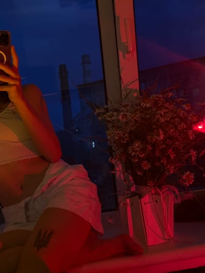 A person is sitting on a windowsill, holding a phone. They are wearing a white top and white shorts. A tattoo is visible on their left thigh. In front of them is a vase with a bouquet of white daisies, lit by red light. Outside the window, the sky is dark blue, and two industrial chimneys are visible. The scene is illuminated by a red light source, possibly a lamp or colored lighting. The image appears to have been taken at dusk or night.