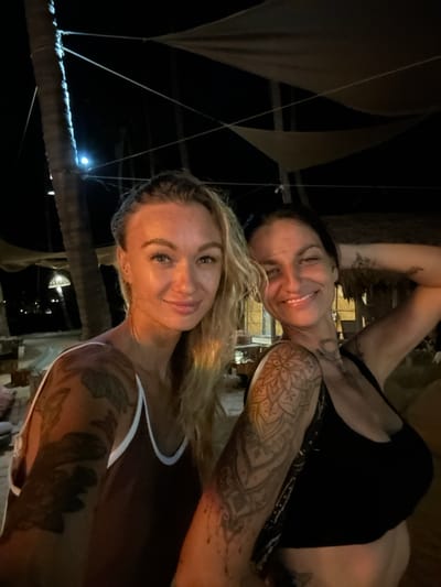 The image depicts two women posing for a selfie at night in Mui Ne, Vietnam. The woman on the left has blonde hair and is wearing a dark brown top with white trim. She has numerous tattoos on her arms and torso. The woman on the right has dark hair and is wearing a black top. She also has tattoos on her arms and torso. They are standing outdoors, with palm trees and a canopy structure visible in the background. Lights are present in the distance, suggesting a resort or beachfront area. The overall atmosphere is casual and relaxed.