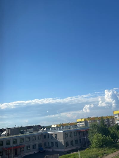 This media file presents a bright, sunny daytime scene of a residential area in Марковский, Russia. The upper portion of the image is dominated by a clear, vivid blue sky dotted with scattered white clouds. Some clouds are fluffy and cumulus-like, while others form long, thin streaks, particularly in the middle-right section of the sky.
Along the bottom of the frame, a row of multi-story apartment buildings is visible. On the left side, buildings feature distinctive yellow accents, possibly on balconies or upper facades, contrasting with their lighter walls. Towards the right, the buildings are predominantly grey with numerous windows. Lush green trees are interspersed among the structures, and patches of green grass are visible at ground level, suggesting common areas or yards. A utility pole or lamppost can be seen on the right, and a small, white, goal-like structure is partially visible on the grass further right.
The time of day appears to be either morning or afternoon, given the bright sunlight and clear conditions. No people are visible, and no specific activities are taking place, presenting a tranquil view of an urban residential landscape. There is no visible text in the image.