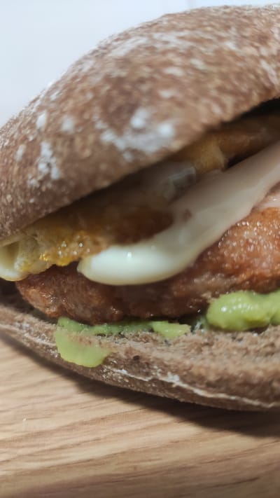 A close-up, slightly blurred shot shows a sandwich made with a dark, rustic bun. The sandwich filling appears to include a thick, browned patty, a slice of melted white cheese, and a layer of green, mashed avocado. The sandwich is resting on a light brown, wooden surface. The background is out of focus and appears to be a white or very light-colored surface. The image does not contain any information about its location in Madrid, Spain.