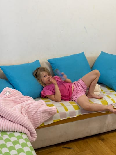 A young child with blonde hair, wearing a pink t-shirt and pink shorts, is lying upside down on a couch. The couch is adorned with colorful pillows, including blue, yellow, and white checkered ones. A pink, fuzzy blanket is also draped over the couch. The child is holding a blue toy in their hand and appears to be looking at it. The setting seems to be indoors, with a white wall to the left and wooden flooring visible on the right. The overall scene suggests a casual, relaxed moment, possibly playtime or a moment of rest. There is no visible text in the image.