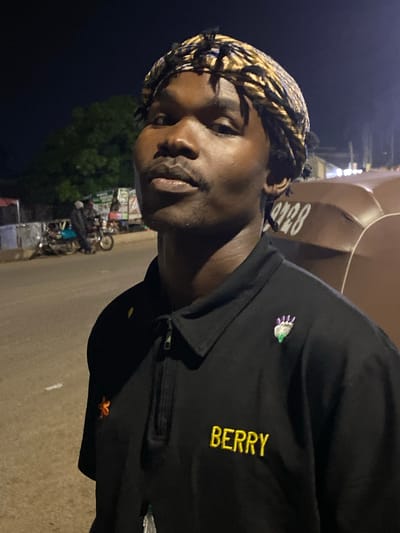 This is a close-up, horizontally oriented photograph of a young person with dark skin and braided hair. They are looking towards the camera with a neutral expression. The person is wearing a black collared shirt with a zipper closure. The shirt has two embroidered details: a small purple and green cluster of shapes on the left chest area and the word "BERRY" embroidered in yellow on the right side of the chest.
The background is blurred and dark, suggesting it is nighttime. A large, curved, brown object, possibly part of a vehicle or structure, is visible behind the subject. In the distance, there appears to be a street scene with some trees and what looks like a motorcycle and various market stalls or roadside vendors. This hints at an outdoor setting, likely a public area at night. The overall lighting suggests artificial illumination, possibly from streetlights or the vendors' setups. The image has a slightly grainy quality, common in low-light photography.
The location context provided is Bukuru, Nigeria. This information, combined with the visual cues of market stalls and a motorcycle, suggests the photograph was taken in a bustling area of Bukuru, likely during an evening or night market. There is no clear indication of the weather conditions. The emotion conveyed by the subject is one of calm observation.
