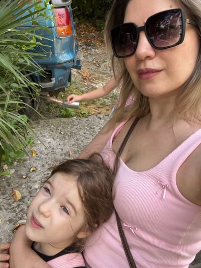 This is a close-up, selfie-style photograph taken outdoors. A woman wearing large, dark sunglasses and a pink tank top is in the foreground, with a young child resting on her shoulder in the lower left quadrant. The woman has blonde hair and is looking at the camera. The child has brown hair and is looking upwards and slightly to the left, with a pensive expression.
In the background, a partially visible blue car with a damaged rear bumper is parked. A child's hand is visible in the mid-ground, reaching towards the car's exhaust pipe. Lush green foliage is present on the left side of the frame, obscuring part of the car and the ground. The ground appears to be a mix of concrete and gravel, with scattered fallen leaves. The overall lighting suggests it is daytime, possibly sunny, given the shadows.
The location is the city of Sochi, Russia, specifically on a pathway or roadside, indicated by the presence of vegetation, a car, and the textured ground surface. The environment appears somewhat unkempt, with the car showing signs of disrepair and the ground covered in leaves.