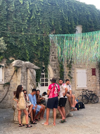 A group of young adults are gathered outdoors, appearing to be taking a break or socializing. The scene is set against a stone building with an ivy-covered wall. In the background, a bicycle is parked, suggesting an urban or historical setting. Above the people, a decorative element made of green netting and shimmering, iridescent strands hangs, possibly part of a temporary installation or decoration.
The individuals are casually dressed, some in t-shirts and shorts, others in dresses and light clothing, indicating warm weather. They are seated and reclining on what appears to be a textured stone surface or steps. Some are looking at each other, engaged in conversation, while others are looking away, perhaps at their phones or in different directions. The overall mood seems relaxed and informal.
A small sign with text is visible on the stone wall. While the text is partially obscured and not entirely clear, it seems to read "The Square of Poets." This, along with the architecture and the general ambiance, hints at a location in a historic town or a tourist area, possibly in Budva, Montenegro, as suggested by the location context. The lighting suggests it is daytime, likely late morning or afternoon, with bright sunlight casting shadows.