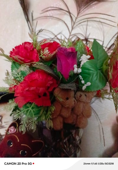 A close-up, slightly unfocused, indoor shot features a bouquet of artificial red roses and a single pink rose, interspersed with green artificial leaves and small white floral accents. Two small, brown teddy bear figures are positioned in the lower center, partially obscured by the flowers. To the left, a dark red object with large black eyes, resembling a stylized animal face, is visible. The background is a textured, light-colored wall with a subtle sparkle effect. The image displays camera information at the bottom: "CAMON 20 Pro 5G" and "24mm f/1.65 1/20s ISO3218." The scene appears to be a decorative arrangement within a home or indoor setting. There is no information in the image to determine a specific location in Yakutsk, Russia.
