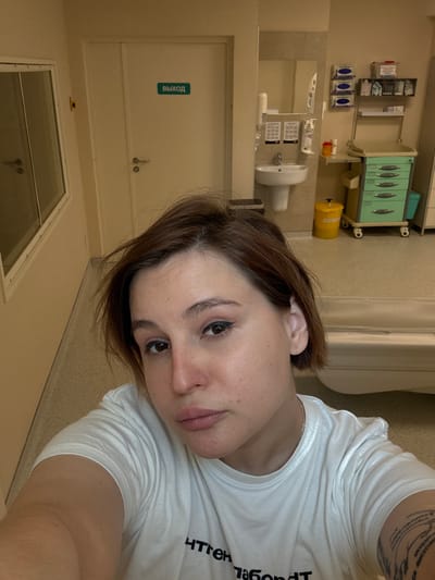 A young woman is captured in a selfie, taken at a downward angle, likely from a lying or reclined position. She has short, dark brown hair and is looking towards the camera with a neutral to slightly pensive expression. Her eyes are made up with eyeliner. She wears a white short-sleeved t-shirt, and a silver chain is visible around her neck. A tattoo with abstract designs is visible on her left upper arm/shoulder.

The background suggests an institutional or medical setting, likely a hospital or clinic in Odintsovo, Russia, given the visible text. The walls are a light, neutral color. On the wall to the left, there are several wall-mounted dispensers and holders containing what appear to be medical supplies like gloves or wipes. Below these is a sink with a mirror, and further right, a yellow cylindrical container, possibly a biohazard bin, on a wheeled cart. Above this cart is a light green medical cabinet with multiple compartments.

In the lower left portion of the image, a white door is visible, featuring a blue sign that reads "ВЫХОД" (VYKHOD), which is Russian for "Exit." Below the door, there's a window or glass panel, possibly looking into an adjacent room or hallway. The partially visible, inverted text on the woman's t-shirt appears to read "ТЕН ЛАБОРАТОР", likely part of "РЕНТГЕН ЛАБОРАТОРИЯ" (X-ray Laboratory), further confirming the medical environment.
