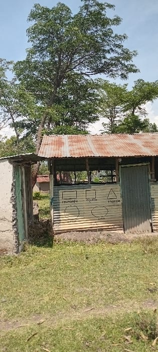The image displays an outdoor scene in Kisumu, Kenya, under a clear blue sky. The central subject is a single-story, open-sided structure constructed with corrugated metal sheets for walls and a heavily rusted corrugated metal roof. The front exterior wall of this structure features hand-drawn geometric shapes: a rectangle, square, triangle, circle, oval, and hexagon, each labeled with its English name in black lettering. A closed corrugated metal door is visible on the right side of the structure. To the left, a partial concrete or masonry wall, with some green paint residue, connects to the main structure via a smaller, also rusted, corrugated metal lean-to roof section. The foreground consists of green grass interspersed with patches of exposed soil and scattered dried leaves. In the background, numerous large trees with dense green foliage are visible, and a small building with a reddish-brown roof is partially discernible behind the main structure. No individuals are present in the scene.