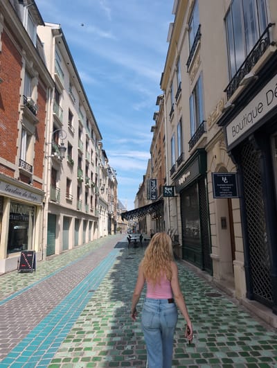 A woman with blonde, wavy hair, wearing a pink tank top and light blue jeans, walks away from the camera down a narrow cobblestone street in Reims, France. The cobblestones have a distinct pattern of grey, green, and blue tiles. On the left side of the street, a brick building has signs for "Gardien du Temps" and "ANTIQUITES". Across from it, on the right, are shops with awnings and signs, including one that reads "Boutique Décoration". The street is lined with multi-story buildings, some with wrought-iron balconies and shuttered windows. The sky above is blue with scattered clouds. In the distance, a tall tower, possibly part of a cathedral, is visible.