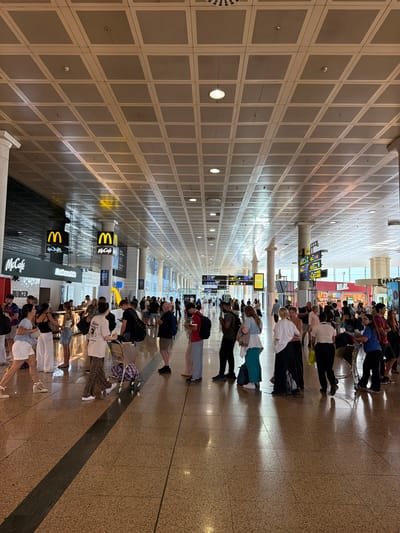 The image depicts a long, wide hallway in what appears to be an airport terminal. The ceiling is a grid of square panels, with linear lighting fixtures casting a warm glow. The floor is tiled with a speckled pattern. On either side of the hallway, people are seated and standing, suggesting they are waiting. Many are holding phones or luggage.
Visible text includes "REINX" on a red sign and the McDonald's logo with "McCafé" on it. These indicate the presence of commercial establishments within the terminal. The overall atmosphere seems to be one of calm waiting, with people engaged in their own activities. The lighting suggests it could be daytime, possibly mid-morning or afternoon, given the brightness in the distance. The presence of an airport setting is further confirmed by the directional signs visible in the background, with yellow and blue signage common in transportation hubs. The architecture, with its clean lines and repetitive patterns, is typical of modern airport design.