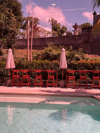 This is an outdoor scene in Kuta Selatan, Indonesia, featuring a swimming pool and a seating area. The swimming pool occupies the foreground, filled with clear, light blue water, reflecting shadows of palm fronds. Along the edge of the pool, a paved area is visible, with a concrete border. Behind this, a wooden bar-style table is set up with several red wooden chairs. To the left of the table, a closed, light pink umbrella stands on a concrete base. Behind the seating area, a dense green hedge forms a backdrop, with some dry, reddish foliage interspersed. Further back, a stone wall is visible, topped by more greenery and some trees. In the distance, a traditional Balinese pagoda structure can be seen above the wall. Two tall, thin wooden poles stand vertically in the mid-ground, with a faint contrail stretching across the pinkish-purple sky. The overall lighting suggests it is daytime, possibly late afternoon or early morning, given the color of the sky and the presence of strong shadows. There are no people visible in the image.