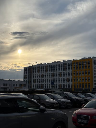 The image depicts a street scene in Kazan, Russia, characterized by modern apartment buildings and a row of parked cars. The buildings are tall, multi-story structures with a mix of white and bright yellow facades, featuring numerous windows. The cars are parked parallel to the buildings, with their fronts facing the viewer or slightly to the side. Visible car brands and models include an Audi with a license plate reading "E026PA 716". A parking sign is also visible, indicating a parking area.

The time of day appears to be either sunrise or sunset, with the sun low in the sky, casting a warm glow through the clouds. The sky is filled with dramatic, wispy clouds, suggesting dynamic weather conditions, possibly indicative of an overcast or partly cloudy sky. The overall mood is somewhat serene, with the setting sun creating a soft, diffused light. The presence of residential buildings and parked cars suggests an urban or suburban environment. No people are clearly visible in the image. The overall impression is that of a quiet moment in a contemporary urban landscape.