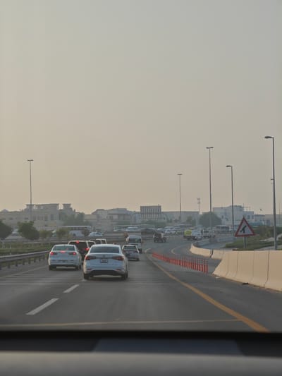 The image depicts a view from inside a vehicle on a highway in Dubai, United Arab Emirates. The sky is overcast with a hazy, light beige color, suggesting either a dusty atmosphere or an early morning or late afternoon time of day. Numerous cars are visible on the multi-lane highway, some of which appear to be in traffic. A white sedan with license plate "15838" is in the foreground, followed by other vehicles. On the right side of the road, a beige concrete barrier lines the highway, with a series of orange and white traffic cones in front of it. A triangular road sign with a black arrow indicating a lane split is also visible. In the distance, buildings with distinctive architectural features, possibly wind towers, are visible, hinting at traditional Middle Eastern design elements integrated into modern structures. Streetlights are interspersed along the highway. The overall impression is one of a typical day on a busy urban highway, likely during a period of moderate traffic, under a diffused, hazy light.