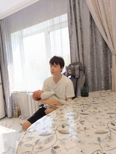 A fair-skinned young male with short, dark hair is seated indoors, holding a fair-skinned infant wrapped in a white blanket. He wears a white t-shirt and dark shorts. They are positioned at a floral-patterned table covered in a tablecloth. To the right of the young male is a grey gaming chair with a headrest featuring a logo. On the table, beside the chair, is a green glass. A window with sheer, light-colored curtains stands behind them, allowing bright daylight into the room. Heavy, grey, patterned curtains frame the right side of the window. To the left, behind the window, is a jar on the floor. The location is Andreyevo, Russia.