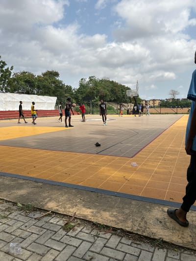 This outdoor daytime scene, captured in Lagos, Nigeria, features a bustling basketball court under a cloudy sky. The court itself is distinct, with alternating sections of yellow and grey textured flooring, delineated by red lines.

Numerous individuals, mostly young males, are present on the court, engaged in what appears to be a basketball practice or game. In the mid-ground, a person wearing a black jersey with "BROOKLYN NETS" clearly visible on the front stands holding a basketball. Other players are scattered across the court; one in a red shirt with a basketball, and another in a yellow shirt. A basketball hoop with a backboard is visible in the background on the left side of the court, where more people are gathered.

The foreground shows the lower body and foot of a person, wearing dark slip-on shoes, standing on a paved concrete area adjacent to the court. This paved area is composed of grey interlocking bricks and concrete, with some dry leaves and sparse grass. The court is surrounded by lush green trees and shrubs, and a tall white banner or tent structure is visible behind the court on the left. In the further background, a metal tower and some buildings can be seen under the bright, overcast sky. The overall atmosphere suggests an active community sports event or training session.