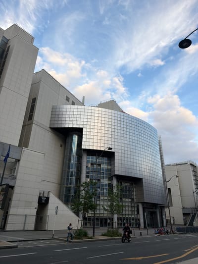 The image shows the Opéra Bastille in Paris, France, taken from street level. The building is modern, with a reflective, grid-like facade contrasting with the more textured, light-colored stone of the lower levels. The sky is partly cloudy. A few individuals are present on the sidewalk and street. One person is walking and looking at their phone. A person is riding a bicycle. Bicycles are parked on the sidewalk. A street lamp is visible.