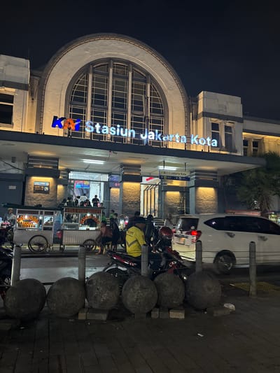 The image depicts the entrance of the Jakarta Kota train station at night, located in West Jakarta, Indonesia. A prominent sign above the entrance reads "KA Stasiun Jakarta Kota" in illuminated blue and orange lettering. The station building features a large arched window and is illuminated by warm lighting. In front of the station, several food carts are visible, with vendors and customers interacting. People are seen standing, walking, and sitting around the carts and near the entrance. Motorcycles are parked in the foreground, and a white car is driving past. The ground is paved with dark tiles, and large, rounded stone objects are placed along the sidewalk. The overall scene suggests a busy public space at night, with commercial activity and transit operations.
