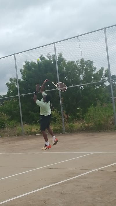 An adult male, wearing a light green short-sleeved shirt, dark shorts, white socks, and orange and beige athletic shoes, is depicted mid-air on a tennis court, performing an overhead tennis stroke. His left arm is extended upwards, and he holds a tennis racket with his right arm, positioned to strike a yellow-green tennis ball visible in the upper left quadrant of the image, near the top of the chain-link fence. The court surface is a hard, light-colored material with white boundary lines. Behind the player, a chain-link fence with metal posts stretches horizontally, separating the court from an overgrown area with numerous green trees and sparse vegetation. The sky is overcast and grey. This scene is located in Lokoja, Nigeria.