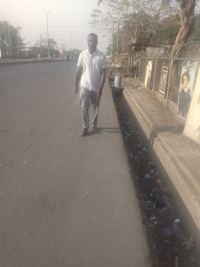 The image shows a man walking along a paved road in New Karu, Nigeria. He is wearing a striped shirt, grey trousers and sandals. He is walking on the right side of the road, towards the camera, with a concrete curb and gutter to his right. There is litter in the gutter. The road surface is grey and there is a concrete sidewalk to the right of the road. There are buildings and trees on the right side of the road. The image is taken from a street level perspective. The weather is clear and sunny.
