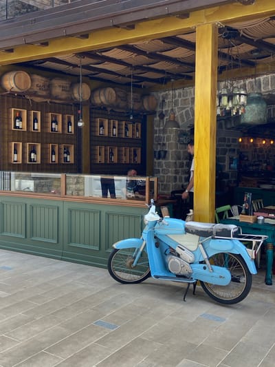 The image shows a blue vintage moped parked on a stone-tiled sidewalk in front of a building. The building appears to be a cafe or restaurant with a rustic aesthetic. The facade features a green wooden lower section and a glass display case. Above the display case, shelves are stocked with wine bottles and burlap sacks labeled "CAFE BRASIL." Several wooden barrels are also visible on a shelf above the sacks. The upper part of the building has a yellow-painted structural beam and a thatched ceiling. Behind the moped and to the right, there are tables and chairs set up, with a person standing near a yellow pillar. The overall scene suggests an outdoor seating area in a town, possibly in a Mediterranean region given the architectural style. The image is taken during the daytime.