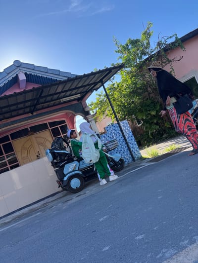 In Choeng Thale, Thailand, a light blue electric scooter is parked on the side of a paved road, near a house. Two children, a boy wearing a white cap and green tracksuit and a girl with a white hijab and green tracksuit, are seated on the scooter. The boy is looking away from the camera, while the girl is looking forward. Both have backpacks on. A woman, wearing a black head covering, a patterned black and red outfit, and carrying a black shoulder bag, stands to the right of the scooter, facing the children. She is gesturing with her right hand. The house behind them has a pink facade, a covered porch with a metal roof, and decorative blue and white tiles. Lush green foliage is visible around the house. The sky is clear blue with some wispy clouds.