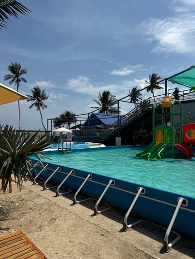 This is a medium shot of a water park on a sunny day, likely located in Mũi Né, Vietnam. The foreground is dominated by a bright blue, above-ground swimming pool filled with clear turquoise water. White metal supports hold up the sides of the pool, and a wooden lounge chair is visible in the lower left corner. To the left of the pool, a large, spiky green plant and a yellow umbrella stand out against the sandy ground and the ocean in the distance.
In the middle ground, a children's water play structure with slides in green and red is situated within the pool. A person is visible on a platform above the structure, wearing a dark outfit and a hat. Stairs lead up to this platform, and a large blue-roofed building with a darker metal frame is visible behind it.
The background features several tall palm trees swaying gently, suggesting a tropical coastal location. The sky is a vibrant blue with scattered white clouds. The overall scene conveys a sense of leisure and recreation.