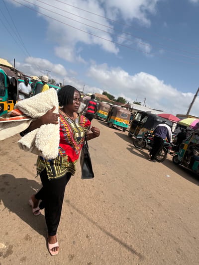 The image shows a woman in a vibrant, patterned dashiki and black pants walking on a dusty road. She carries a fluffy white blanket in her arms, and appears to be walking away from the camera. She is wearing light pink slip-on sandals. The scene is a bustling street with several three-wheeled vehicles, known as tricycles or "keke napeps," parked and in motion. Some of these vehicles are brightly colored, with yellow, green, and blue accents. In the background, more people are visible, as well as market stalls with colorful awnings. The sky is blue with scattered white clouds, suggesting it is daytime. The overall atmosphere appears to be that of a busy marketplace or transport hub. The image is oriented vertically, with the sky occupying the upper portion and the ground the lower. The light indicates it is likely midday. There is no visible text in the image.
