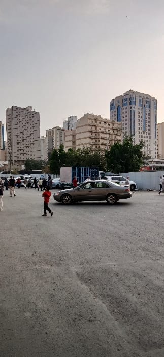 This image depicts an outdoor urban environment in Makkah Al Mukarramah, Saudi Arabia. The foreground features a large expanse of unpaved, grey ground. A light brown sedan is centrally positioned, facing slightly left, either parked or moving slowly. A young individual wearing a red shirt and dark pants walks leftward in front of the sedan. To the left and behind the vehicle, a group of approximately 15-20 pedestrians, comprising adults and possibly children, are visible, predominantly moving towards the left. Their attire includes both casual clothing and traditional Middle Eastern garments. In the mid-ground, behind the pedestrians and sedan, additional vehicles are present, along with green trees and a prominent blue metal container or structure. The background consists of numerous multi-story buildings, including a tall, light-colored structure with many square windows on the far left, several tan-colored buildings with balconies in the center, and a distinct modern high-rise with blue-tinted windows and white architectural elements on the far right. The sky is a uniform light grey, indicating an overcast or hazy condition.