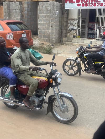 In an urban setting in Abuja, Nigeria, two individuals are riding on a red motorcycle. A man, appearing to be the driver, is dressed in a light green traditional outfit and is seated at the front. Behind him, a woman wearing a black shirt and blue pants is the passenger. The motorcycle has a visible engine and a silver exhaust pipe. In the background, an orange car is parked next to a building constructed with grey bricks. The building has a sign with red lettering that reads "MEDICINE" and a phone number. Another motorcycle, driven by a person in a checkered shirt and red cap, is visible to the right, moving in the same direction. The ground is a paved road.