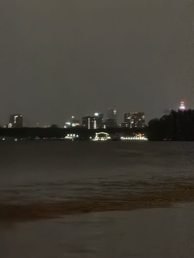 The image captures a nighttime view of a cityscape, likely Moscow, Russia, across a body of water. The skyline features several illuminated buildings of varying heights, with some appearing to be skyscrapers. There are also what seem to be boats or barges with lights on the water in the foreground, and trees or a dark shoreline separating the water from the buildings. The overall atmosphere is dim, suggesting it is nighttime, possibly with some fog or low cloud cover obscuring the sky. The water surface shows gentle ripples. The city is identified as Khimki, Russia.