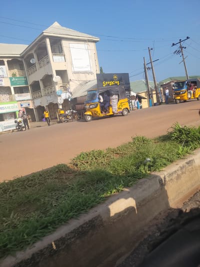 The image depicts a street scene in Lokoja, Nigeria, on a bright, sunny day. The perspective is from inside a vehicle, looking out at a bustling street. Several yellow and blue auto-rickshaws, commonly known as "keke napep" in Nigeria, are visible. Some are carrying passengers and goods, suggesting ongoing transportation activities.
One prominent auto-rickshaw has "Spagking" written on its side in bold yellow letters. Other auto-rickshaws have license plates that read "AJ71A" and "AJ3348". In the background, a multi-story building with a corrugated metal roof is visible. Signs on the building include "JENISTEPH MADE & MAKE-UP STUDIO" and "RISMEDIX".
There are also people on the street, some walking, some standing near the auto-rickshaws. Overhead, electrical wires crisscross the scene, supported by wooden poles. The overall impression is one of everyday life and commerce in a Nigerian city. The lighting suggests it is daytime, likely morning or afternoon, with clear skies.