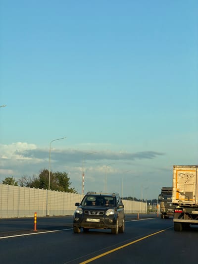 An eye-level, full shot captures a highway scene in Kaipy, Russia, under a clear, pale blue sky. The road is asphalt, marked with yellow and white lines. In the foreground, a black SUV drives in the right lane. The SUV has its headlights on, and its license plate reads "B 150 PC 716". Inside the SUV, two people are visible. To the right of the SUV, a large semi-truck with a white trailer is in the adjacent lane. The trailer has an orange and white graphic on its side. Further down the road, another semi-truck is visible. On the left side of the highway, a sound barrier wall runs parallel to the road, punctuated by orange traffic cones. Several streetlights are also visible along the highway. Trees and foliage are present behind the sound barrier.