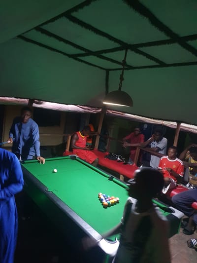 The image captures an indoor night scene in Lokoja, Nigeria, where several young men are gathered around a pool table. The setting appears to be a casual, dimly lit recreational space, possibly constructed with wooden beams and planks, giving it a somewhat informal and rustic feel.
In the foreground, a dark-skinned man wearing a blue long-sleeved shirt is actively engaged in playing pool. He is leaning over the green-felted pool table, holding a cue stick, and appears focused on his shot. A white cue ball is visible near the edge of the table, while a full rack of fifteen billiard balls, comprising solids and stripes, is neatly arranged in a triangle at the opposite end of the table, ready for play.
Several other men are observing the game. To the right of the pool table, part of another person, possibly wearing a blue garment, is visible. In the background and above the pool table, a group of men is situated on what appears to be an elevated platform or mezzanine, some leaning on a railing. Among them, one man wears a red and white striped jersey, another a light grey t-shirt, and a third a solid red shirt. They are casually watching the game unfold.
A bell-shaped light fixture hangs from the ceiling, casting a warm, bright glow over a portion of the pool table and the surrounding area, indicating it is nighttime or late evening. The ceiling above features a distinctive criss-crossing dark pattern against a green surface. No discernible text is visible on clothing, equipment, or in the environment. The overall atmosphere suggests a relaxed social gathering focused on the game of pool.
