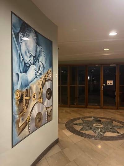 This is a painting displayed in an indoor setting, possibly a hallway or lobby, in Geneva, Switzerland. The painting, framed in dark wood, depicts a man's face and hands, rendered in a blue-toned, realistic style. He appears to be concentrating intently on a complex mechanical object composed of gears and intricate metal parts. The gears are a prominent feature, with some appearing metallic and shiny, while others have a golden hue. The overall scene evokes a sense of precision and craftsmanship, suggesting the subject is a watchmaker or someone engaged in a similar meticulous task.

The surrounding environment shows marble flooring with a decorative starburst mosaic. Behind the painting, there are wooden-framed glass doors, reflecting the interior lights. Two circular lights are visible on the wall above and to the left of the painting. The painting is hung on a plain, light-colored wall. The lighting in the scene appears to be artificial, suggesting it is either daytime with indoor illumination or nighttime. No specific weather conditions are discernible. The artwork conveys a mood of quiet focus and dedication to detail. There is a small sign visible through the glass doors with some text and graphics, but it is illegible in the image.